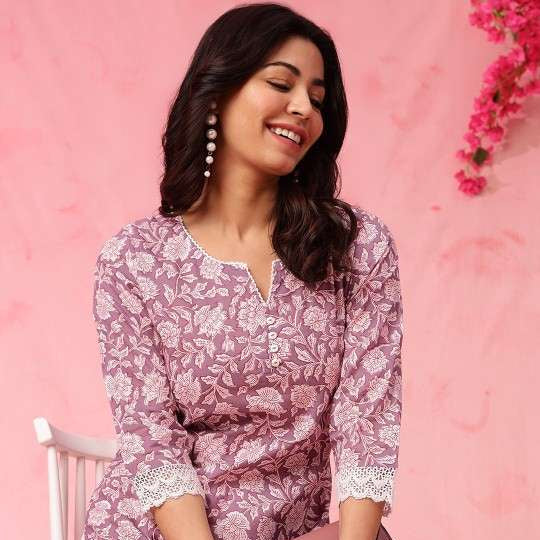 Woman wearing a pink floral top and white pants sitting on a chair with a pink background.